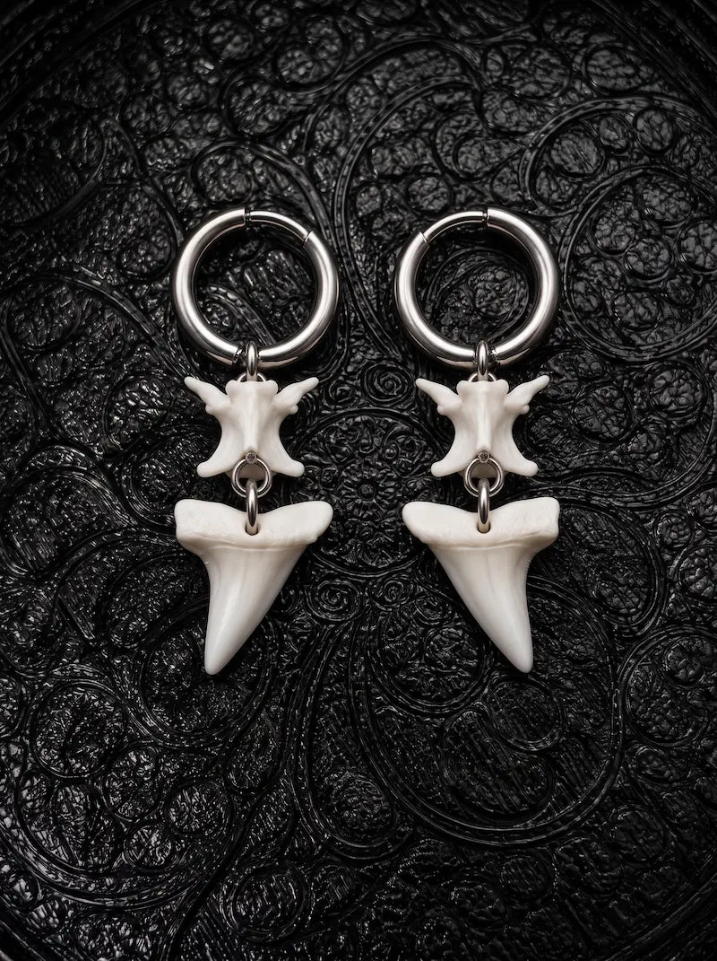 Abyssal mako shark tooth and snake bone earrings with slender pointed teeth, displayed on black patterned fabric