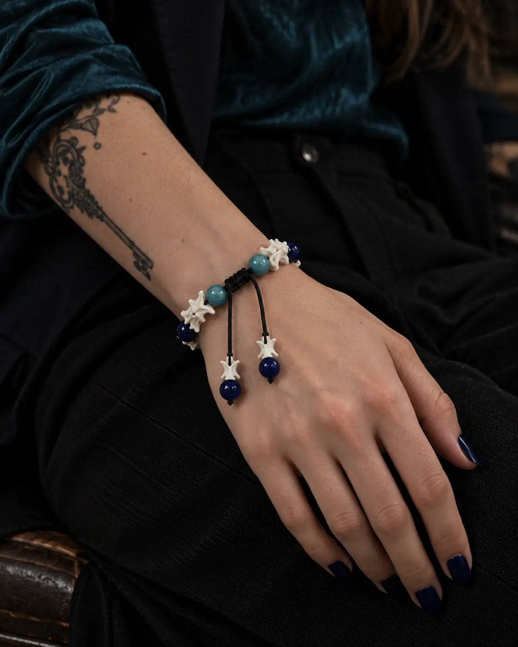 Azure Serpent Eye snake bone bracelet on a hand resting on the thigh, showing two blue bead-tipped cords