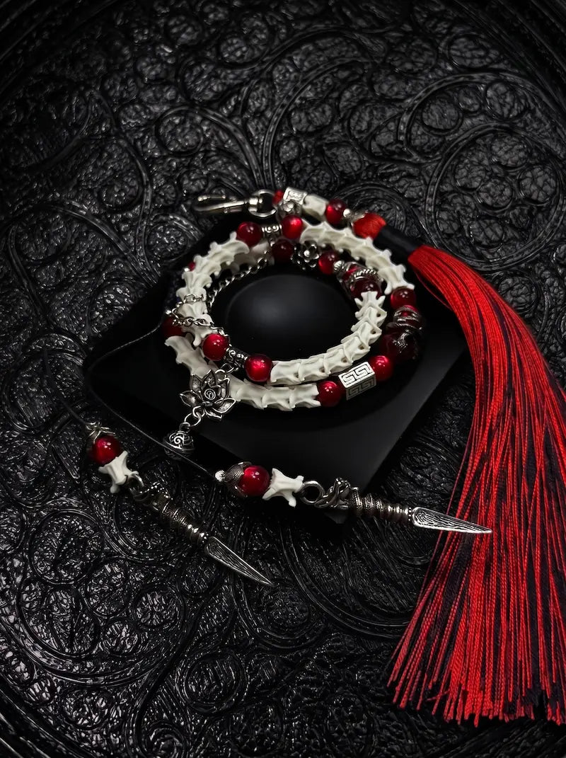 Crimson Lotus double-wrap snake bone bracelet with red beads, a lotus charm and pointed metal drops, beside a red tassel
