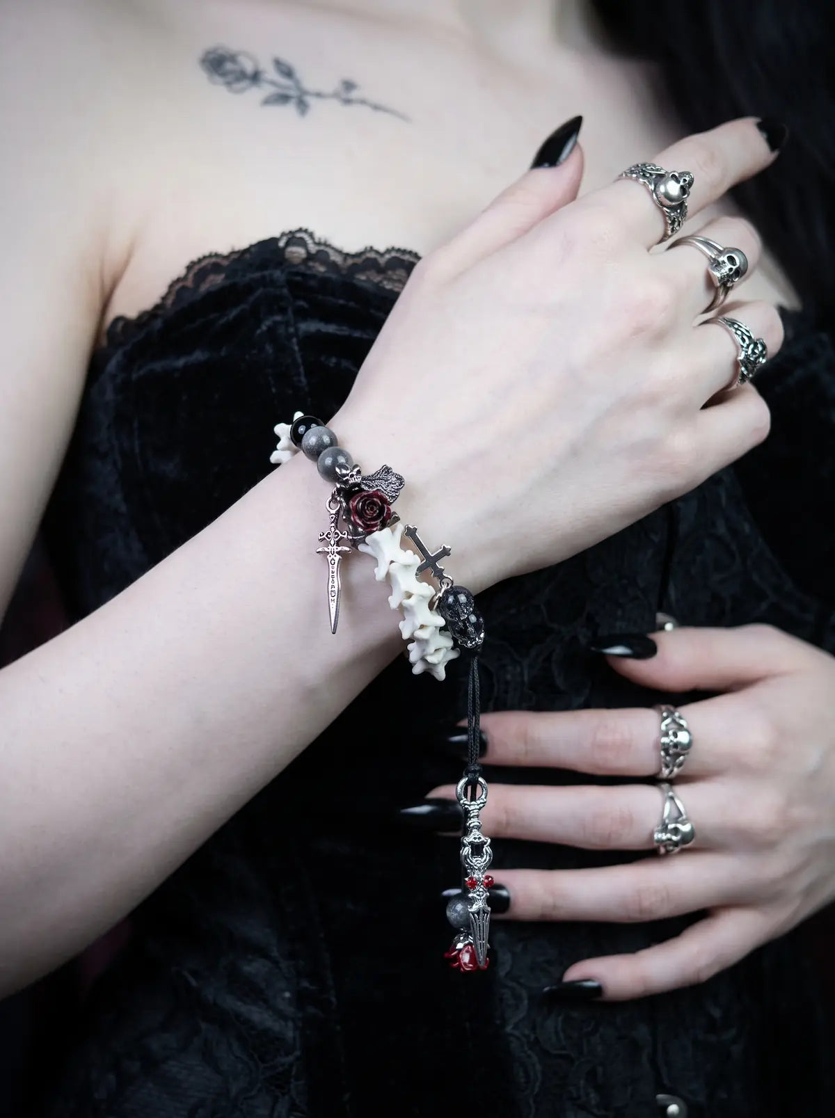 Crimson Rose snake bone bracelet worn with a ringed hand raised above a black corset, with its long cord ends hanging down