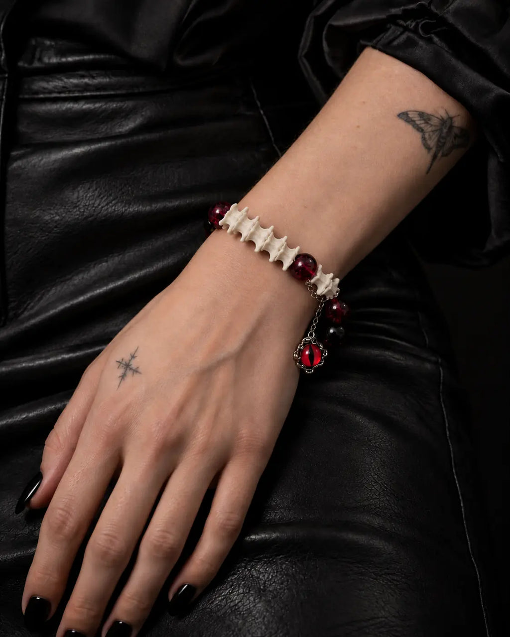 Crimson Serpent Eye snake bone bracelet on a hand resting against black leather, with its red eye charm hanging below