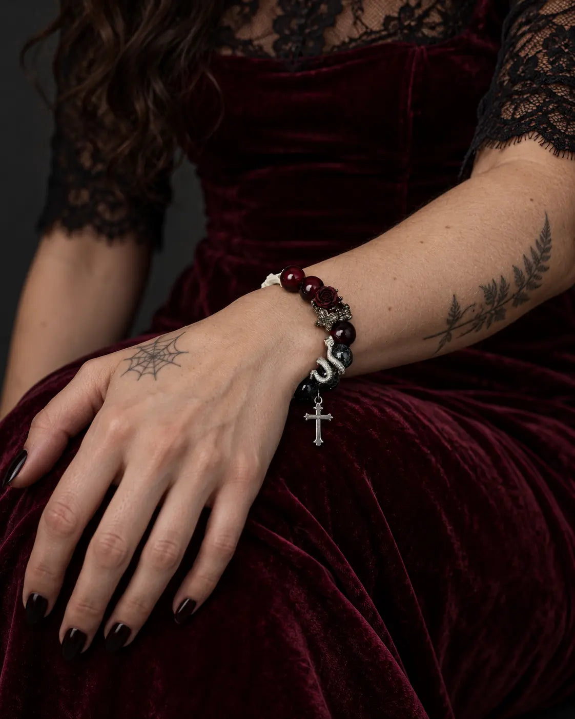Crimson Thorn snake bone bracelet on a hand resting against a burgundy velvet skirt, with its cross hanging below