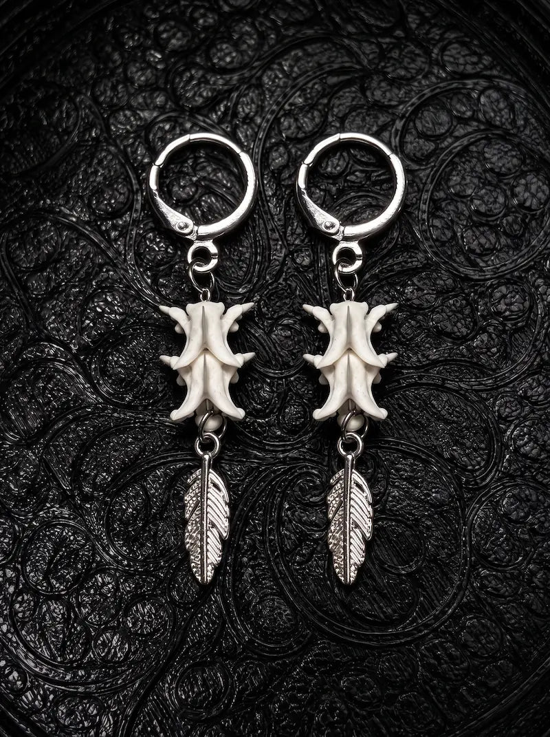 Gravefeather snake bone earrings with engraved silver-tone feather drops, displayed as a pair on black patterned fabric