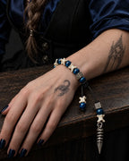 Midnight Relic snake bone bracelet on a hand resting on a wooden table, with its blue beads and pendant visible