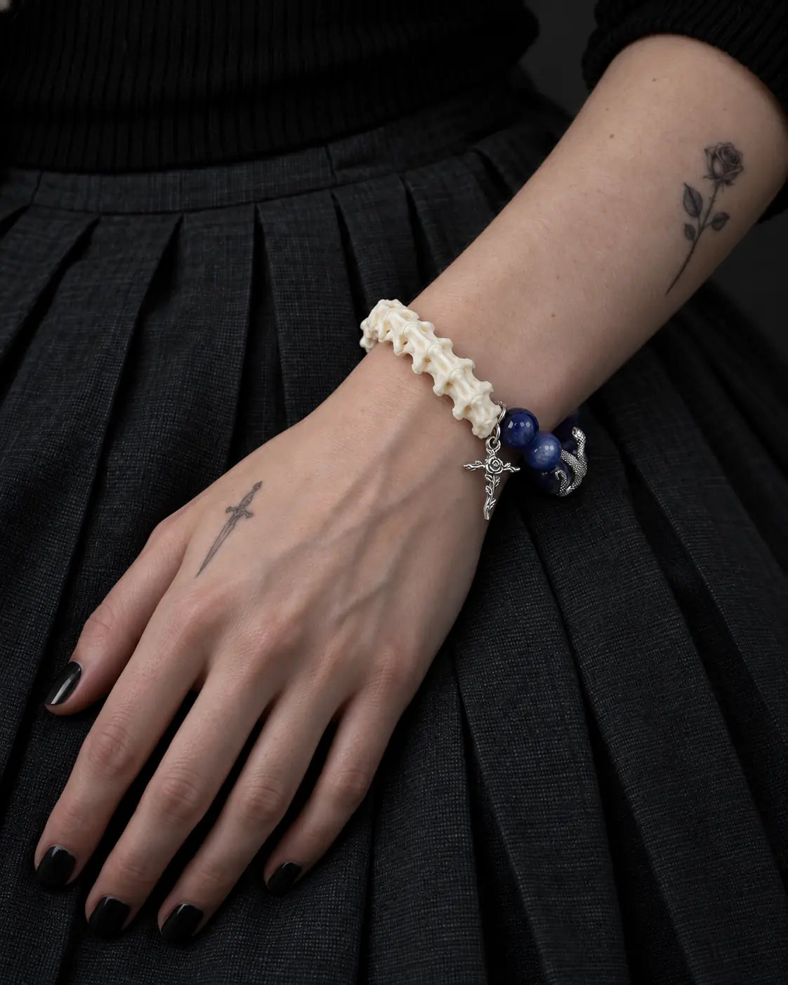 Midnight Serpent Cross snake bone bracelet on a hand resting on a black pleated skirt, with the vertebrae facing upward