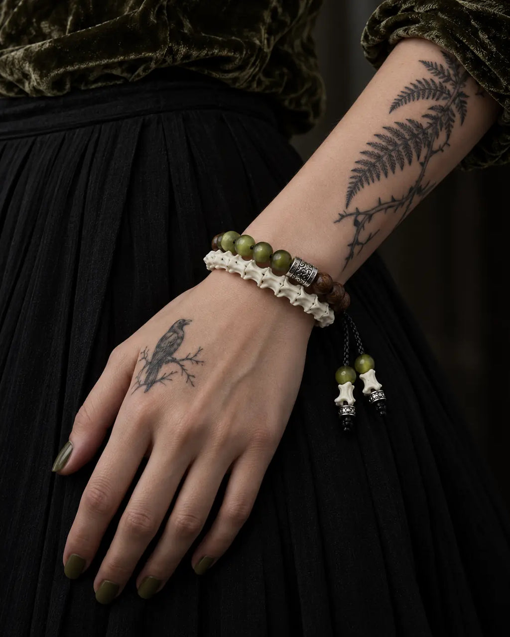 Mosswood snake bone bracelet on a hand resting against a black skirt beneath an olive velvet sleeve