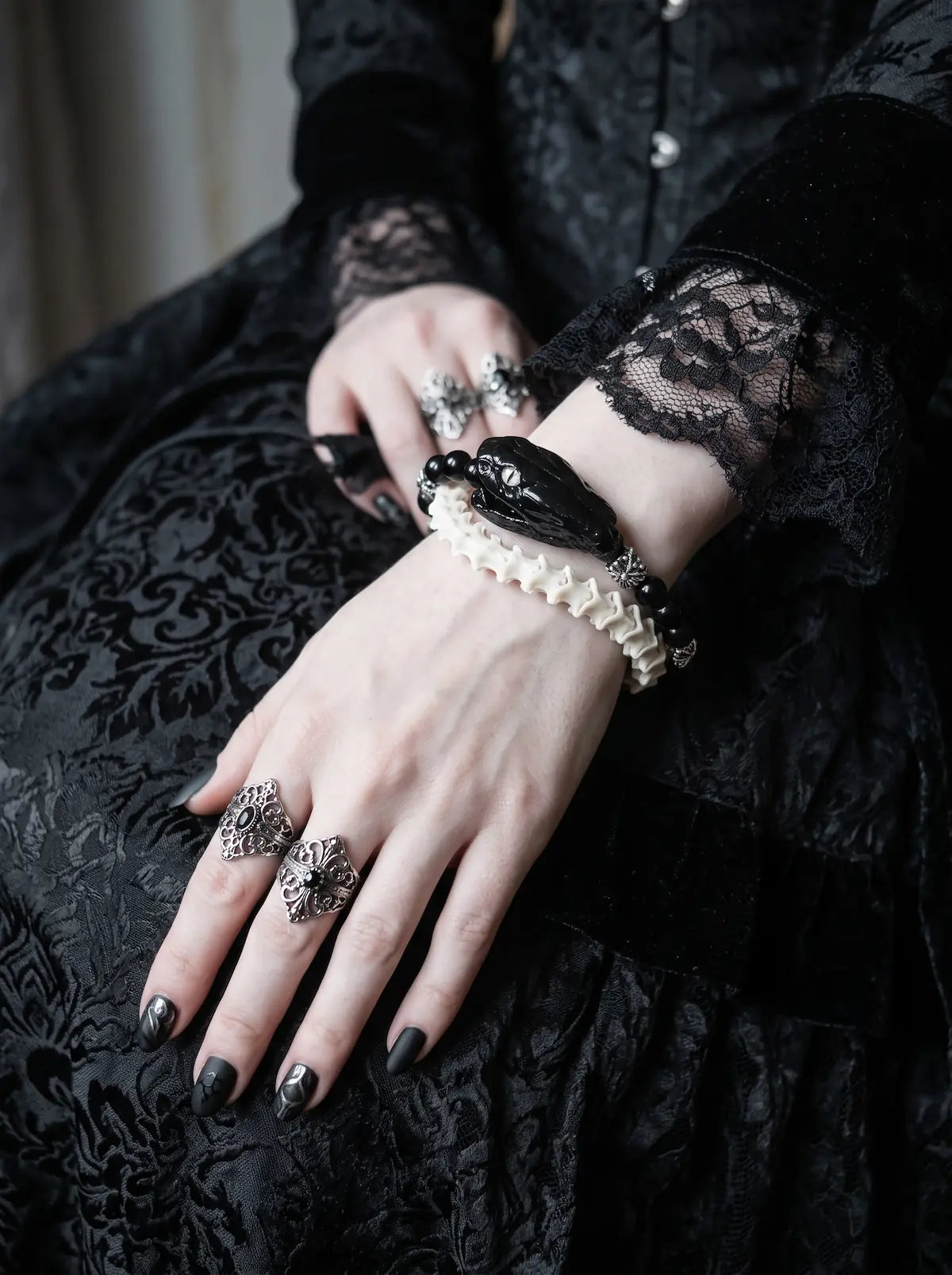 Nightreign Snake Bone Bracelet, wrist detail of pale vertebrae bracelet with dark head and hanging metal dagger