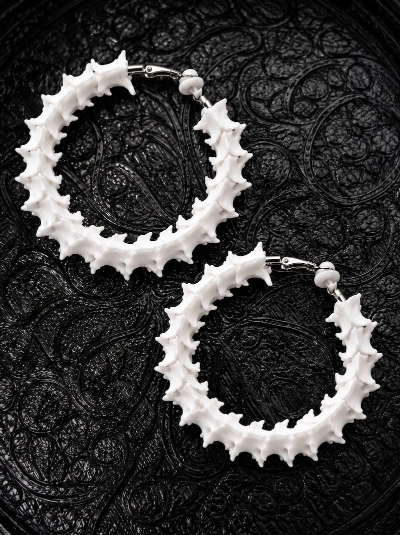 Close-up of Ossuary Hoop snake bone earrings showing white vertebrae around silver-tone hoops on black patterned fabric