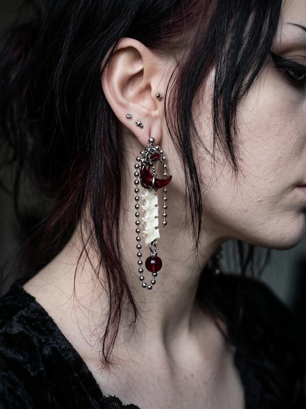 Ravenmoon Snake Bone Earrings, long bone earring worn at the ear, showing red bead and chain accents from the side