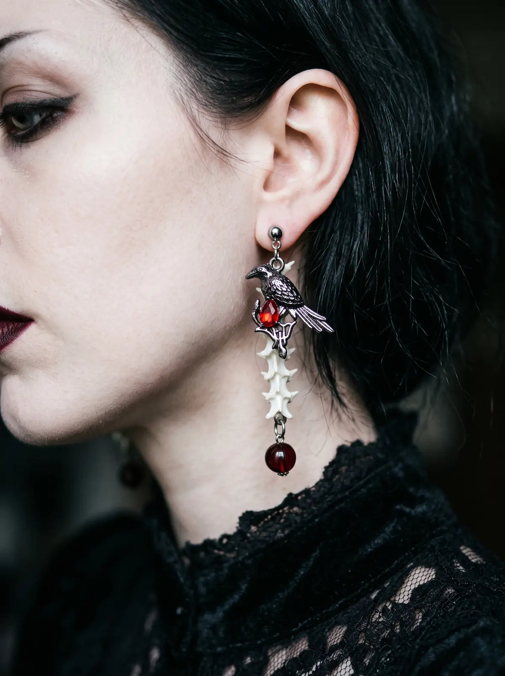 Ravenmoon Snake Bone Earrings, side profile wearing a dangling vertebrae earring with red gemstone-style drops