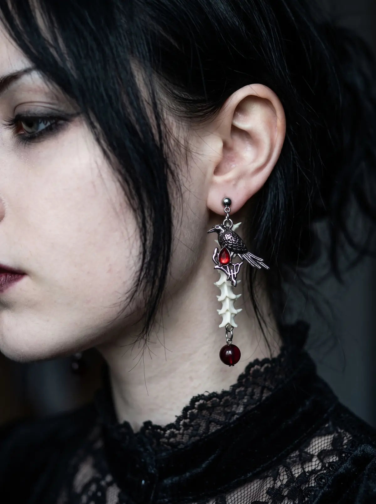 Ravenmoon Snake Bone Earrings, bone-and-red-bead earring hanging beside dark hair in a close side portrait