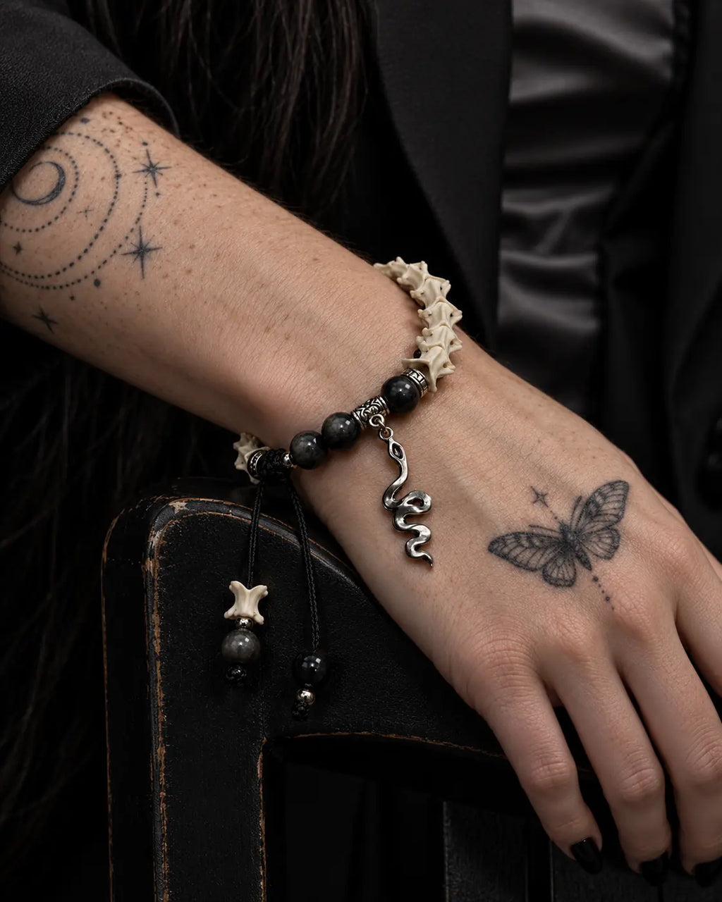 Shadow Serpent snake bone bracelet on a hand resting over a dark chair arm, with its snake charm and cord ends hanging down