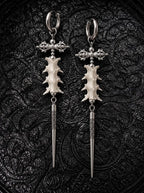 Vajra Spine snake bone earrings with ornate crossbars and long engraved silver-tone spikes on black patterned fabric