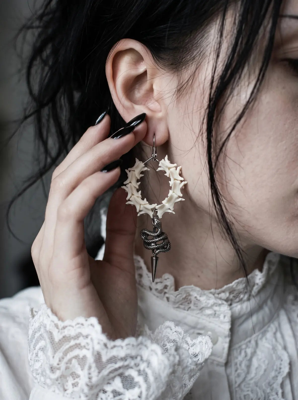 Close-up of a Viper Drop snake bone hoop earring beside a hand with black nails and a white lace cuff